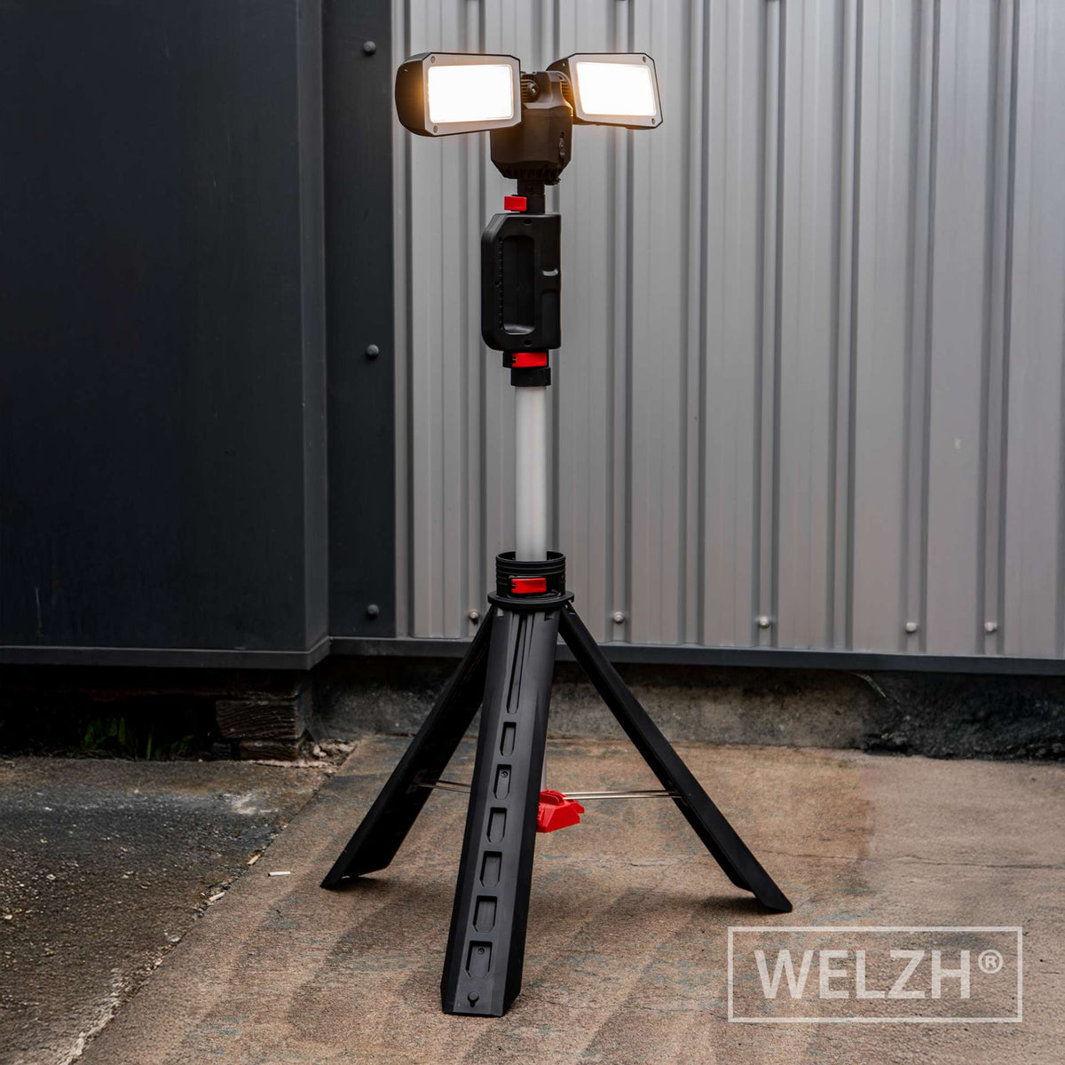 A portable work light on a tripod stand, with two rectangular LED lights illuminated, set against a gray corrugated metal wall and concrete ground. The brand name 'WELZH' is visible as a watermark in the bottom right corner.